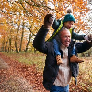 Grandfather Giving Grandson Ride On Shoulders As They Walk Along Autumn Woodland Path